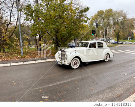 White vintage car driving on wet city road. Retro transport, classic elegance and timeless automotive heritage White vintage car driving on wet city road. Retro transport, classic elegance and timeless automotive heritage 134118313
