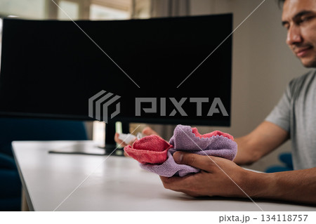 Focused office worker male holding disinfectant spray and microfiber cloth, preparing to clean computer monitor, promoting hygiene and preventing germs in workplace. Concept of healthy lifestyle. 134118757