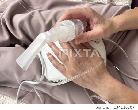 Close up of a portable electric nebulizer inhaler held in the hands of a sick adult woman at home, modern medical device for respiratory therapy, asthma and cold treatment equipment, healthcare 134119207