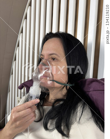 Adult woman using a home nebulizer inhaler while sitting in a cozy bedroom, respiratory treatment at home for breathing relief, healthcare and medical therapy concept, realistic lifestyle scene Adult woman using a home nebulizer inhaler while sitting in a cozy bedroom, respiratory treatment at home for breathing relief, healthcare and medical therapy concept, realistic lifestyle scene 134119210