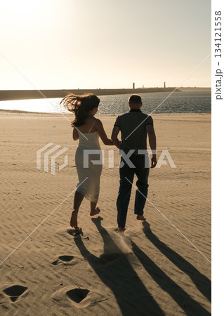 Couple walking barefoot on sandy beach near ocean at sunset, wind blowing her hair in a peaceful romantic scene. Couple walking barefoot on sandy beach near ocean at sunset, wind blowing her hair in a peaceful romantic scene. 134121558