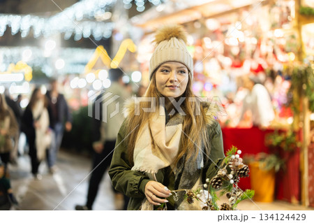 Girl with cone and fir brunch garland stands in middle fair decorated for New Year holidays Girl with cone and fir brunch garland stands in middle fair decorated for New Year holidays 134124429
