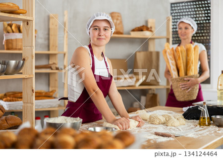 Skilled successful bakers in uniform working in bakery. Young girl working with pieces of dough and adult woman carries basket of baked goods 134124464