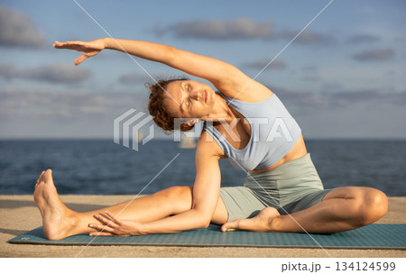Female fitness trainer performing yoga parsvottanasana sitting on mat on beach 134124599