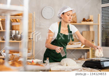 Middle-aged female baker cutting and weighing dough on scales in bakehouse Middle-aged female baker cutting and weighing dough on scales in bakehouse 134124653