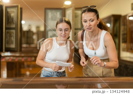 Young woman with teen daughter visitors stands near horizontal glass display case 134124948