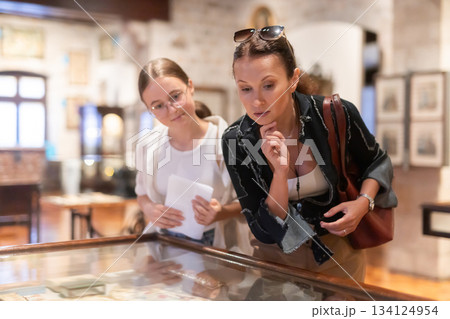Young woman with teen daughter visitors stands near horizontal glass display case Young woman with teen daughter visitors stands near horizontal glass display case 134124954