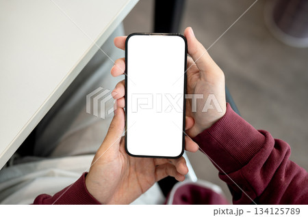 A man holding white screen smartphone with both hands sitting indoors at table in a cafe or library. A man holding white screen smartphone with both hands sitting indoors at table in a cafe or library. 134125789