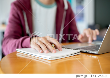 Man student's hand holding pen over notebook and typing on laptop at wooden table in cafe or library 134125828