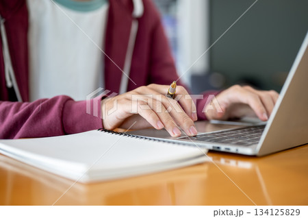 A man student's hands holding pen typing on laptop aside notebook at wooden table in cafe or library 134125829
