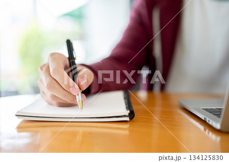 Male student's hand holding pen writing in notebook aside laptop on wooden table in cafe or library. Male student's hand holding pen writing in notebook aside laptop on wooden table in cafe or library. 134125830