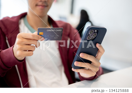 Close up of man holding smartphone looking at credit card sitting indoors at table in cafe or office 134125888