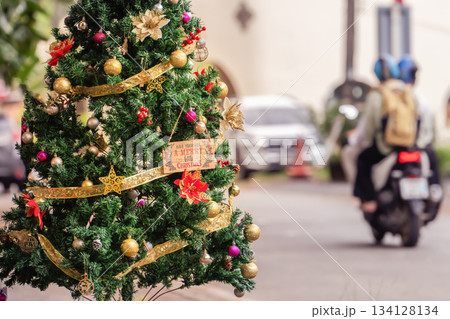 Christmas tree decorated with balloons and a yellow shiny ribbon with sequins on a Phuket street during the day in Thailand, near the road where vehicles and motorcycles drive. High quality photo Christmas tree decorated with balloons and a yellow shiny ribbon with sequins on a Phuket street during the day in Thailand, near the road where vehicles and motorcycles drive. High quality photo 134128134