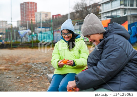 Two Russian women 50 and 60 years old sit outdoors in Vladivostok eating tangerine and enjoying calm mindful time together in cool spring weather Two Russian women 50 and 60 years old sit outdoors in Vladivostok eating tangerine and enjoying calm mindful time together in cool spring weather 134129062