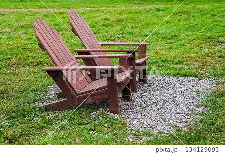 Two wooden Adirondack chairs on a small gravel area in a green grassy field, creating a peaceful outdoor seating background with copy space and nobody around. 134129093
