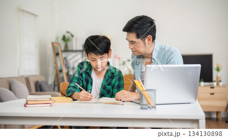 A son writing in the notebook while his father teaching him a homework at their house. A son writing in the notebook while his father teaching him a homework at their house. 134130898