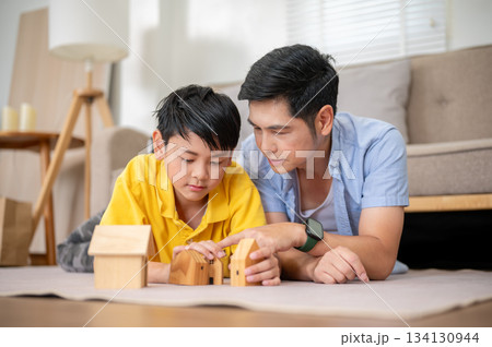 Close up of father teaching elementary son to play with wooden house toy on carpet in living room. 134130944