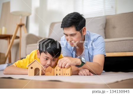 Close up of father and elementary son playing with wooden house blocks toy on carpet in living room. 134130945