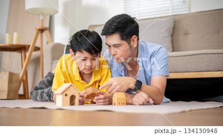 Asian father explaining wooden house toy to his elementary son while laying on living room carpet. 134130948