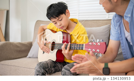 Asian kid boy playing guitar on sofa in living room or music school and father or teacher clapping 134130953