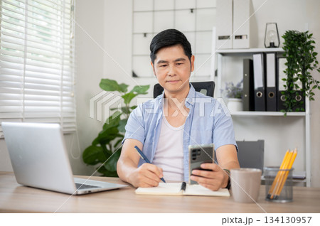 Asian man holding phone and writing with pen in notebook at wooden table in office or working room. Asian man holding phone and writing with pen in notebook at wooden table in office or working room. 134130957