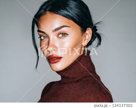 Woman with dark hair and striking features wearing a burgundy sweater poses against a neutral background 134137302