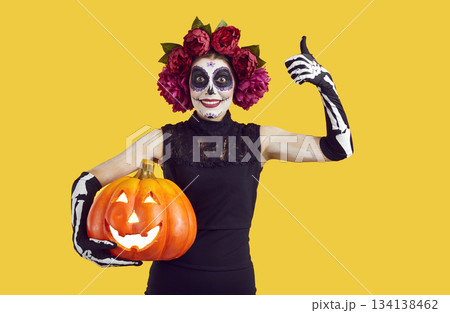 Happy funny Catrina girl holding Halloween jack o lantern, smiling and showing thumbs up 134138462