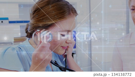 Listening clinician in blue scrubs pressing stethoscope to cheek in exam room, digital overlay 134138772