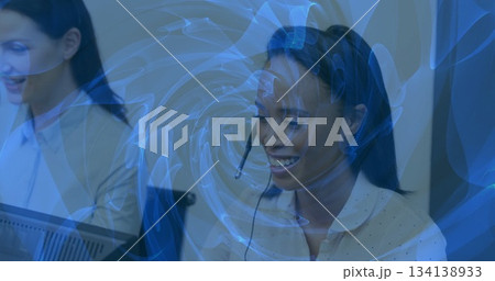 Smiling African American agent wearing blouse and headset handling calls at desks, with blue swirl Smiling African American agent wearing blouse and headset handling calls at desks, with blue swirl 134138933