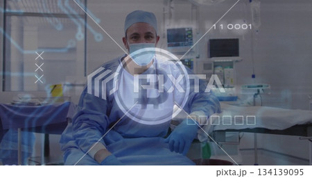 Sitting surgeon wearing blue scrubs and mask in hospital operating room, with Bitcoin logo overlay 134139095