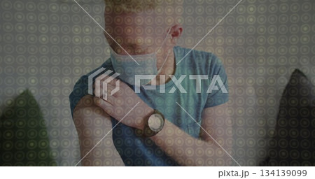 Man wearing protective face mask pulling up shirt sleeve and touching arm at home, with wristwatch Man wearing protective face mask pulling up shirt sleeve and touching arm at home, with wristwatch 134139099