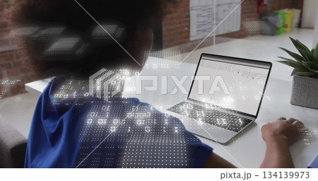Typing woman wearing blue shirt analyzing laptop spreadsheet on office desk, with data overlays 134139973
