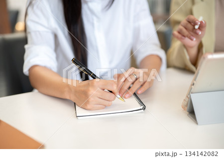 Close up of woman student's hand holding pen writing in notebook with teacher or supervisor talking. 134142082