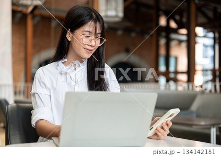 Asian woman student with glasses holding and looking at phone sitting with laptop at table in cafe. Asian woman student with glasses holding and looking at phone sitting with laptop at table in cafe. 134142181