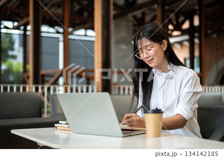 Asian woman student with eyeglasses holding pen writing in notebook over laptop on table in cafe. Asian woman student with eyeglasses holding pen writing in notebook over laptop on table in cafe. 134142185