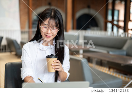 Asian woman student with eyeglasses holding pen and coffee while looking at laptop on table in cafe. Asian woman student with eyeglasses holding pen and coffee while looking at laptop on table in cafe. 134142188