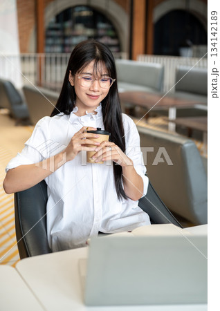 Asian woman student with eyeglasses holding coffee cup while looking at laptop on table in cafe. Asian woman student with eyeglasses holding coffee cup while looking at laptop on table in cafe. 134142189