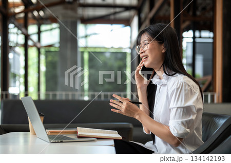 Asian woman student with eyeglasses talking on phone holding pen in front of laptop on table in cafe 134142193