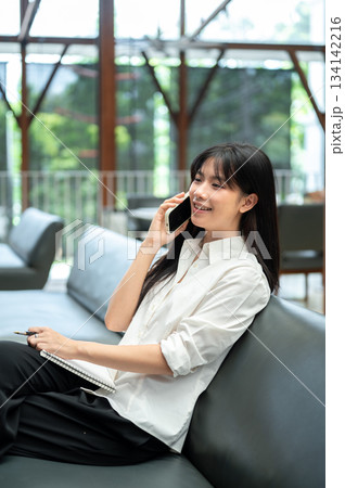 Asian woman freelancer talking on phone and holding pen with notebook while sitting on sofa in cafe 134142216