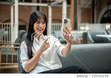 Happy asian woman looking and pointing at phone talking on video call while sitting on sofa in cafe 134142219
