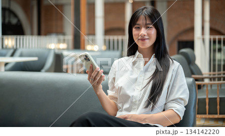 Smiling asian woman holding smartphone as sitting comfortably on leather sofa in cafe or coffee shop 134142220
