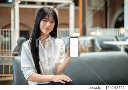 Pretty asian woman holding showing white screen smartphone as sitting on sofa in cafe or coffee shop 134142221