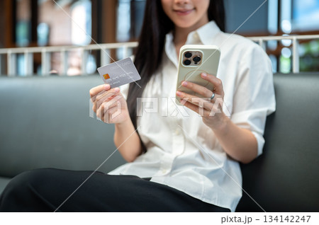 Close up of asian woman holding credit card and looking at smartphone while sitting on sofa in cafe. Close up of asian woman holding credit card and looking at smartphone while sitting on sofa in cafe. 134142247