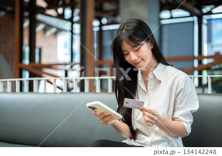 Pretty asian woman holding a credit card and looking at smartphone while sitting on sofa in the cafe Pretty asian woman holding a credit card and looking at smartphone while sitting on sofa in the cafe 134142248
