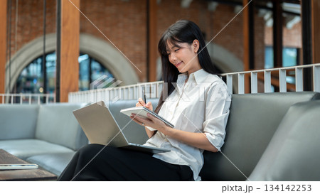 Asian woman student writing in notebook looking at laptop on leg sits on sofa in cafe or coffee shop 134142253