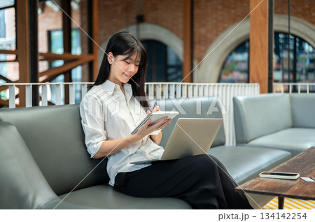 Asian woman student writing in notebook over laptop on legs sitting on sofa in a cafe or coffee shop 134142254