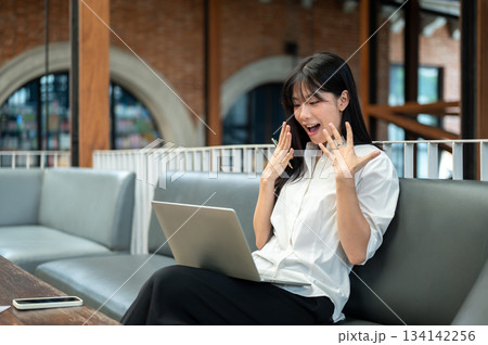 Asian woman student looking at laptop and raising hands in surprised while sitting on sofa in a cafe 134142256