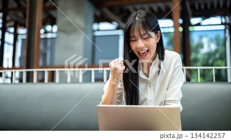 Asian woman student looking at laptop and making a fist in surprised while sitting on sofa in a cafe 134142257