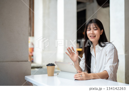 Asian woman student holding tablet and waving hand with pen while sitting at table inside a building 134142263