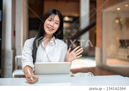 Asian woman student holding tablet and pen as waving a hand while sitting at table inside a building 134142264